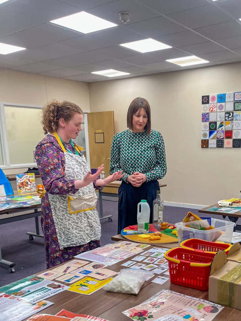 Two women with brown hair stand near each other looking at a display set up. One listens while the other talks